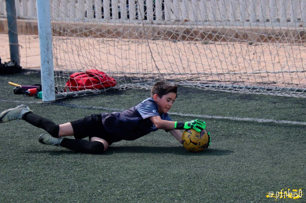 Un portero juvenil atrapando un balón en un campo de césped artificial, mientras se encuentra en el suelo cerca de la portería. Está usando guantes de fútbol y una camiseta deportiva.