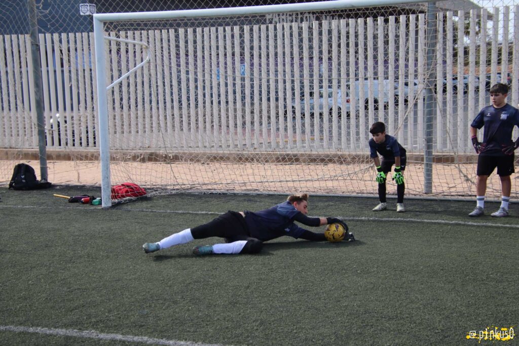 Un grupo de porteros juveniles entrenando en un campo de césped artificial. Un portero está en el suelo atrapando un balón, mientras que otros dos jugadores observan desde la portería, con guantes y ropa deportiva