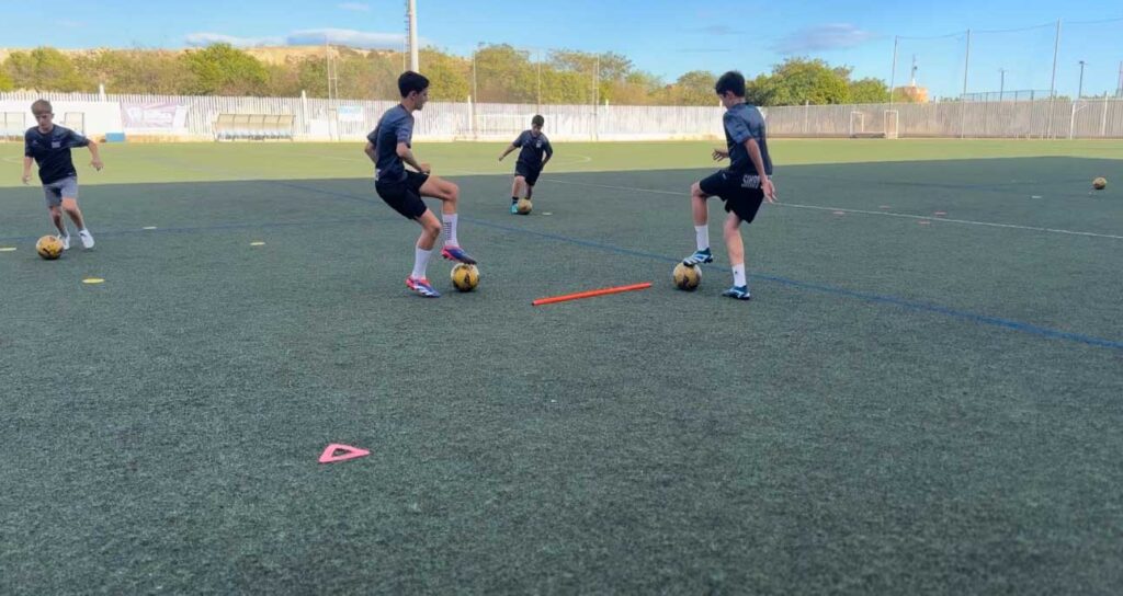 Varios jugadores de fútbol realizando ejercicios de control y pase con balones en un campo de césped artificial. Los jugadores están concentrados en el balón, mientras un par de conos y una barra roja marcan el área de trabajo.