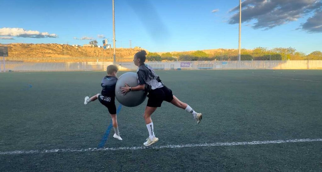 Dos jugadores de fútbol entrenando en un campo de césped artificial. Uno de ellos está sosteniendo una pelota grande mientras el otro la empuja con su cuerpo, ambos vestidos con ropa deportiva de fútbol.